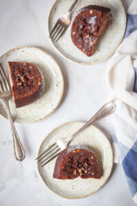 A slice of tunnel of fudge cake on a plate, with the remaining cake behind it on a cake stand.