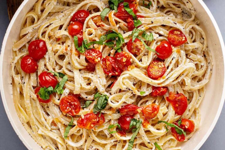 A fork lifts creamy tomato pasta with roasted cherry tomatoes, basil, and black pepper from a white bowl.