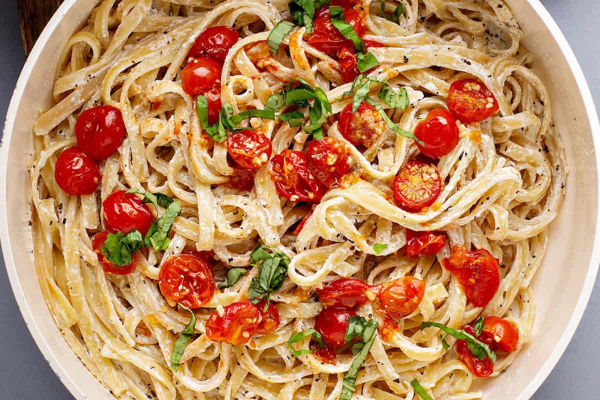 A fork lifts creamy tomato pasta with roasted cherry tomatoes, basil, and black pepper from a white bowl.